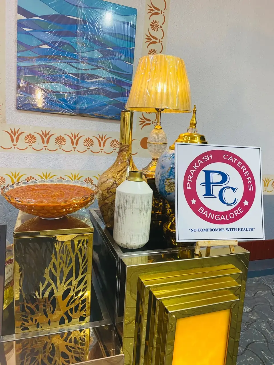 Brass buffet vessels beside a lamp and a Prakash Caterers sign from the previous brand